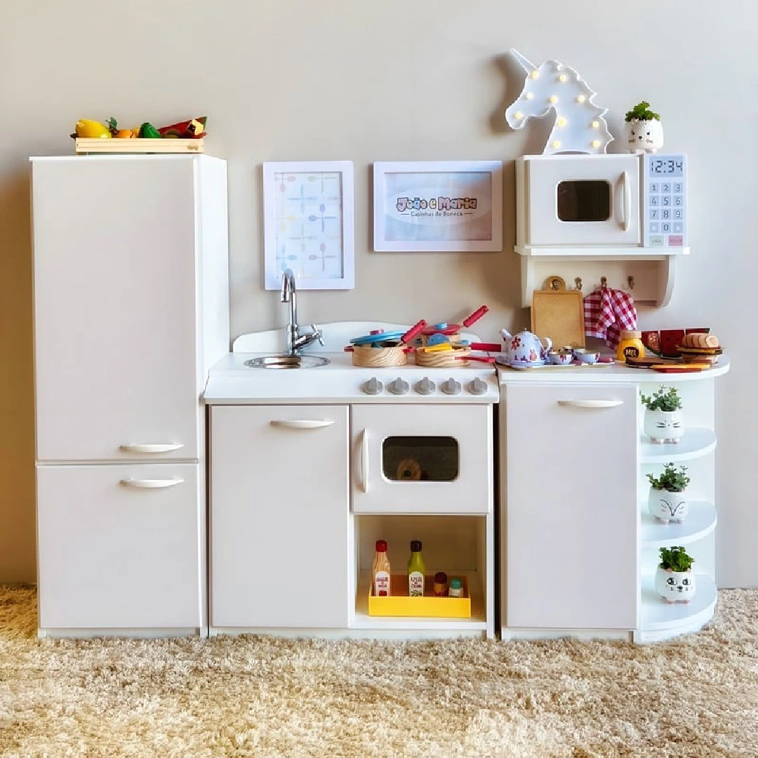 Cozinha infantil branca com geladeira, fogão, pia e micro-ondas, equipada com brinquedos de alimentos e utensílios.