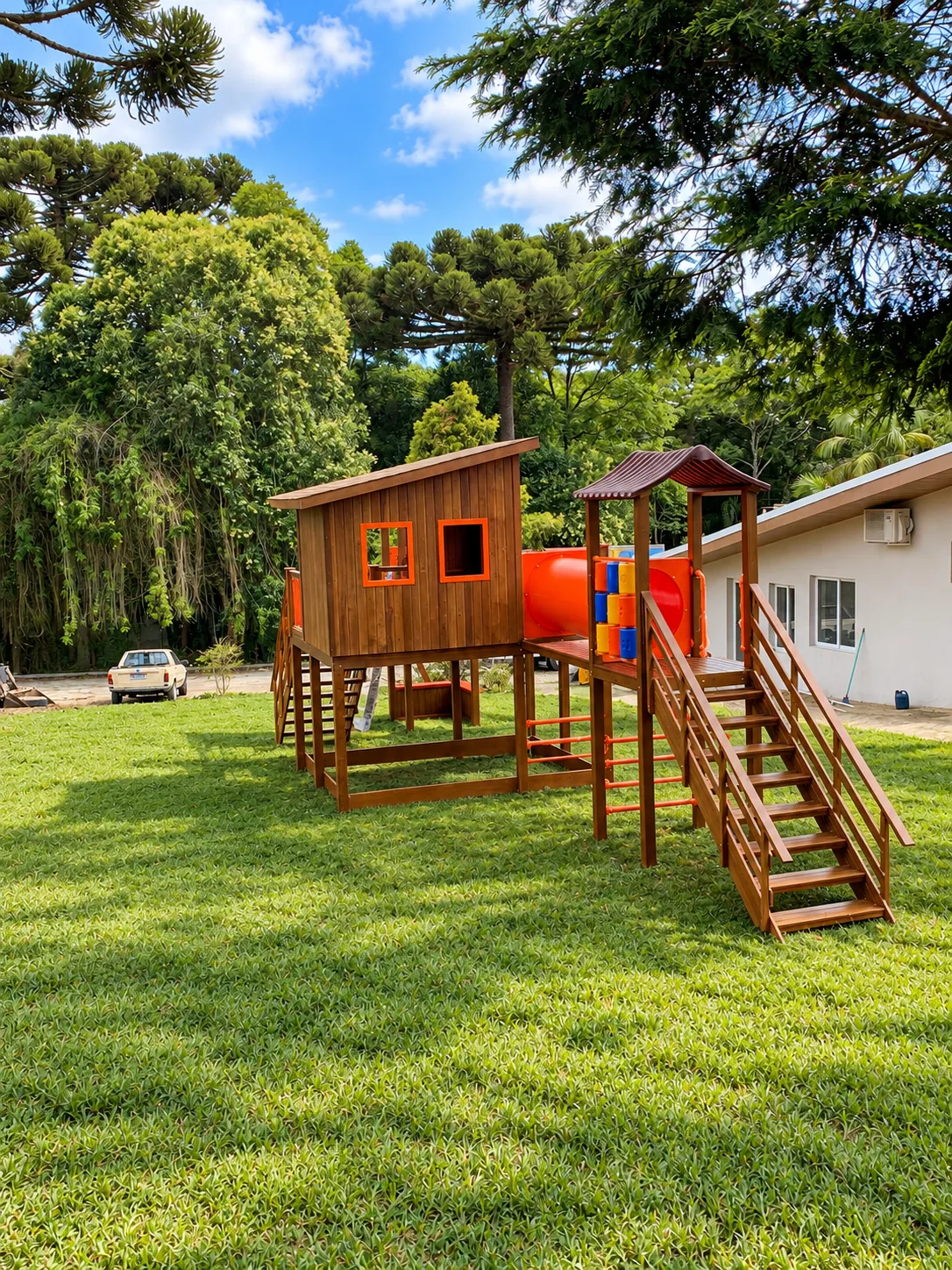 Playground de madeira com torres cobertas e túnel laranja em jardim amplo